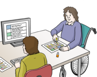 Zwei Personen sitzen zusammen am Schreib-Tisch. Eine Person tippt am PC. Die andere Person liest etwas vor. Sie sitzt in einem Rollstuhl.